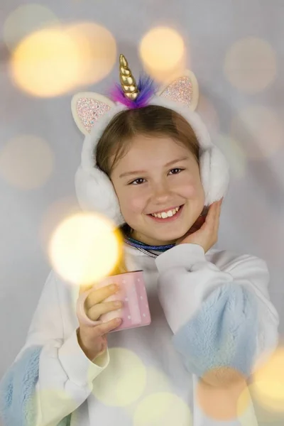 Girl with a headband with a unicorn horn holding a mug. There are lights everywhere.