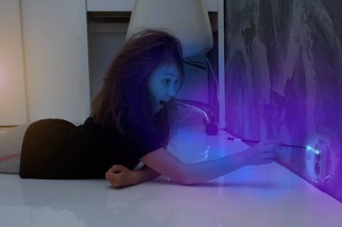 Küçük kız sokete bir asa takıyor. Prizde elektrik akımı var ve kızın saçı dimdik duruyor..