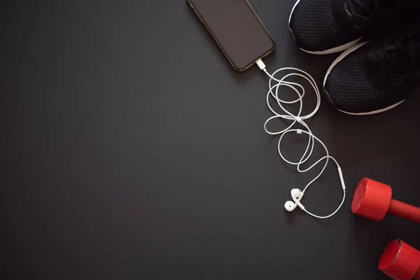 Siyah spor ayakkabılar, spor için kırmızı dambıl ve siyah arka planda kulaklık takan akıllı telefon. Spor malzemeleri seti. Evde eğitim için ideal. Metin için boşluk.