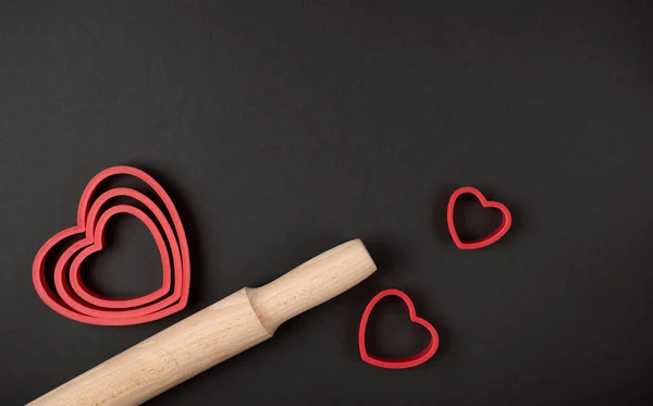 Red heart-shaped baking tins for cookies and wooden rolling pin on the dark (black) background. Flat lay, top view, space for text. Valentine\'s day concept.