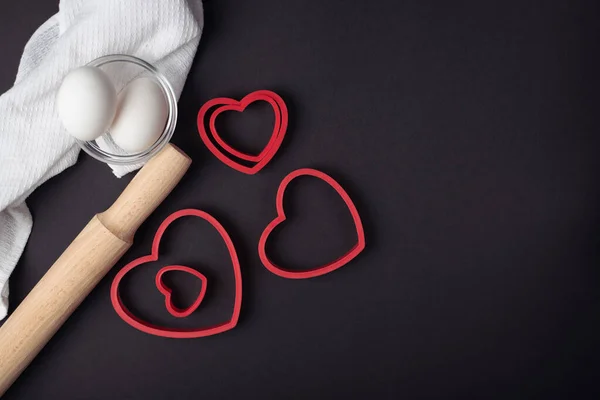 Red heart-shaped baking tins for cookies and wooden rolling pin on the dark (black) background. Flat lay, top view, space for text. Valentine's day concept.