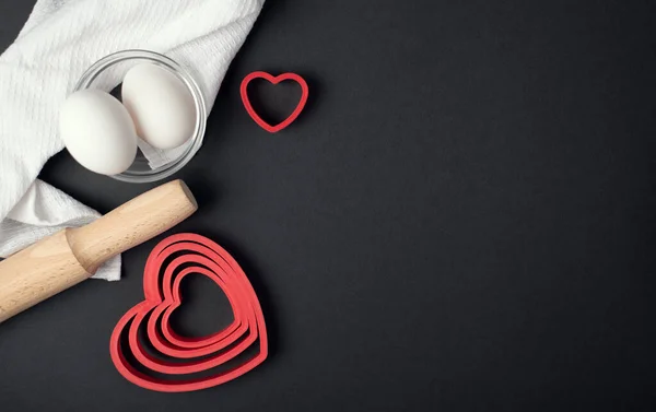 Red heart-shaped baking tins for cookies and wooden rolling pin on the dark (black) background. Flat lay, top view, space for text. Valentine's day concept.