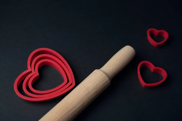 Red heart-shaped baking tins for cookies and wooden rolling pin on the dark (black) background. Flat lay, top view, space for text. Valentine\'s day concept.