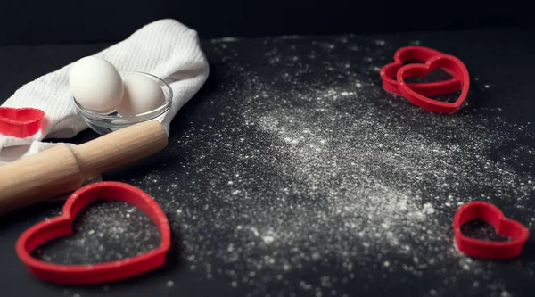 Red heart-shaped baking tins for cookies and wooden rolling pin with scattered flour on the dark (black) background. Flat lay, top view, space for text. Valentine\'s day concept.