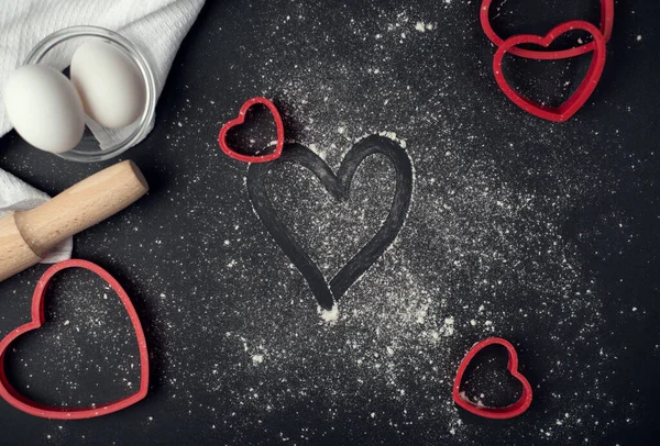 Red heart-shaped baking tins for cookies and wooden rolling pin with scattered flour on the dark (black) background. Flat lay, top view, space for text. Valentine\'s day concept.
