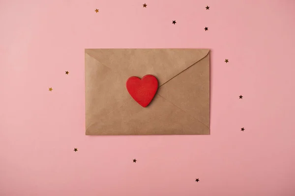 Craft envelope with the red heart and little golden stars on the rose background. Flat lay, top view. Romantic love letter for Valentine\'s day concept.