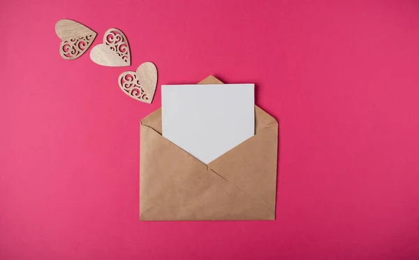 Craft envelope with a blank sheet of paper inside and wooden hearts on the hot pink background. Romantic love letter for the Valentine\'s day concept. Space for text.