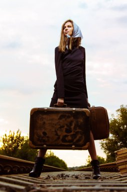 Arka planda iki bavul ve kitapla demiryolunda yürüyen bir kız.