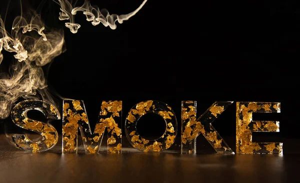 Acrylic letters with gold leaves forming the word smoke with smoke over the letters, on wooden surface, black background, selective focus.