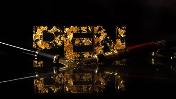 Acrylic letters with gold leaves forming the word pen and pen on reflective surface, black background, selective focus.