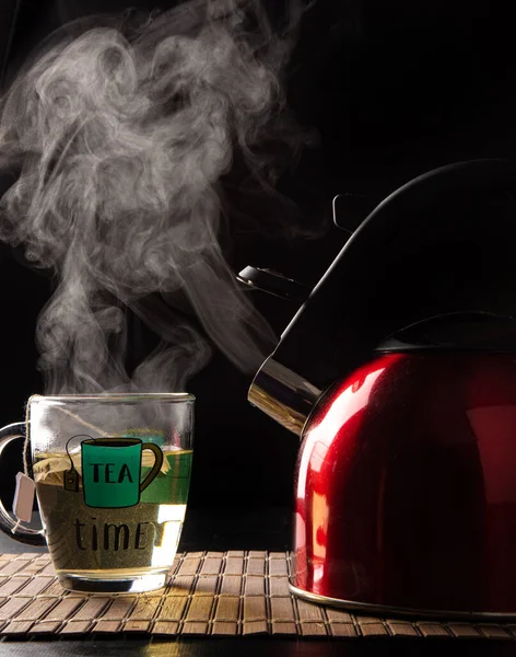 red kettle and cup of tea, both coming out of smoke on a wooden mat, black background, selective focus.