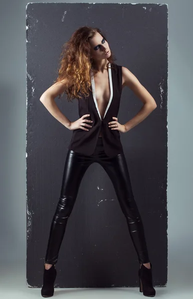 High fashion look of a fashion girl. Portrait of a fashionable model in sexy black leather pants, black boots and beautiful black/white vest. Studio shot. copy space — Stock Photo, Image