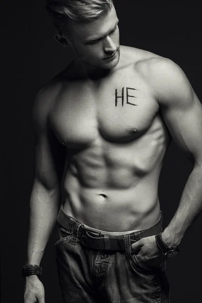 Handsome muscular male model with intense glance posing over grey background. Perfect body with the inscription "he" on his chest . Studio shot — Stock Photo, Image