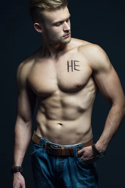 Handsome muscular male model with intense glance posing over grey background. Perfect body with the inscription "he" on his chest . Studio shot — Stock Photo, Image