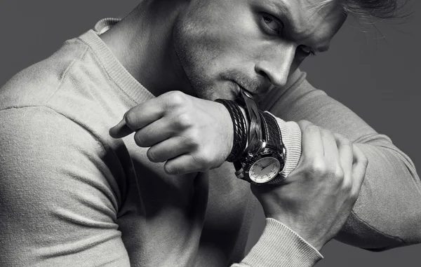 Advertising wrist watch concept. Beautiful (handsome) muscular male model with perfect body in grey jumper. He bites and unfastens the bracelet from the clock. Street style. Black and white, monochrome studio shot — Stock Photo, Image