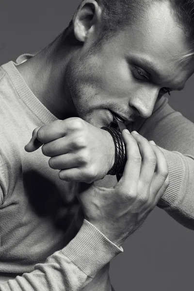 Advertising wrist watch concept. Beautiful (handsome) muscular male model with perfect body in grey jumper. He bites and unfastens the bracelet from the clock. Street style. Black and white, monochrome studio shot — Stock Photo, Image