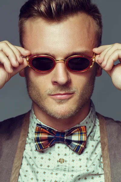 Retrato emotivo de un joven guapo con camisa retro, escocés en pajarita de celda y gafas de sol redondas e inusuales posando sobre fondo gris. Sonrisa blanca brillante y piel sana. Estilo urbano. Captura de estudio — Foto de Stock