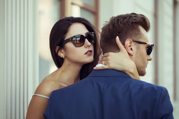 Joven feliz pareja de moda en la ciudad en gafas de sol con estilo. El hombre que lleva en traje de negocios, la mujer en vestido sexy romántico con encaje y abrigo —  Fotos de Stock