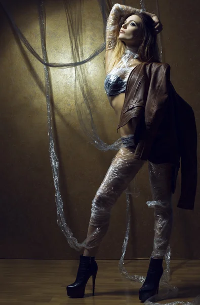 Young brunette sexy woman in a leather jacket and sexy lingerie, wrapped in an oilcloth posing on a gold background. Long legs. Studio shot. The concept of dependence in this world — Stock Photo, Image