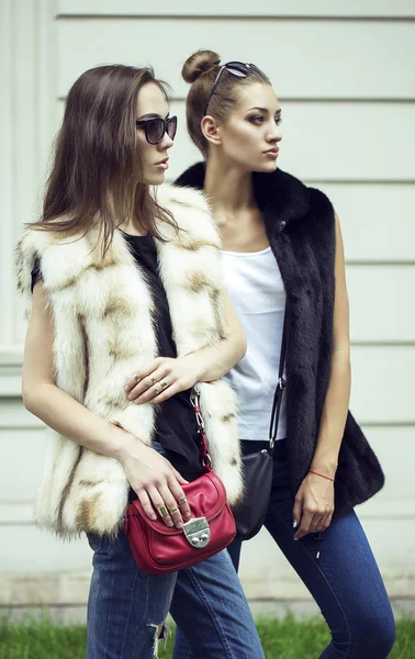 Prise de vue de mode de deux belles filles élégantes dans le coucher du soleil portant des lunettes de soleil, gilets de fourrure. Deux jeunes femmes en plein air dans la rue. Shopping inspiration — Photo