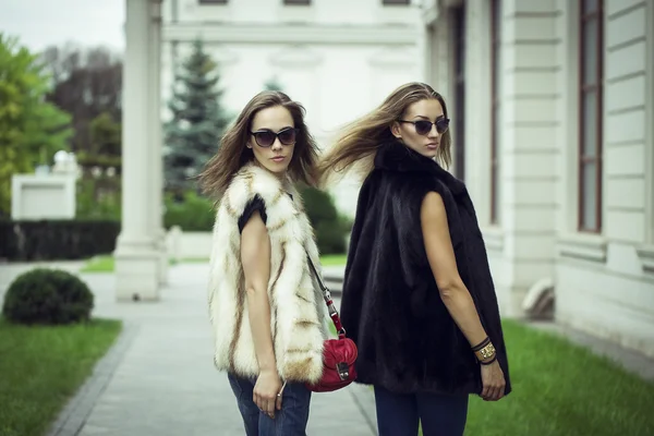 Prise de vue de mode de deux belles filles élégantes dans le coucher du soleil portant des lunettes de soleil, gilets de fourrure. Deux jeunes femmes en plein air dans la rue. Shopping inspiration — Photo