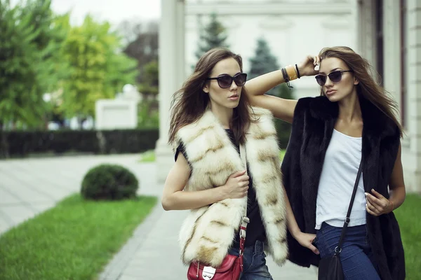 Prise de vue de mode de deux belles filles élégantes dans le coucher du soleil portant des lunettes de soleil, gilets de fourrure. Deux jeunes femmes en plein air dans la rue. Shopping inspiration — Photo