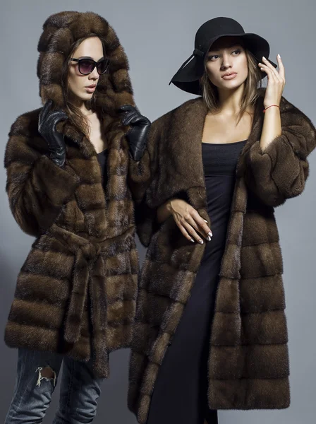 Foto de moda de duas meninas bonitas elegantes (morena e loira) em estúdio em fundo cinza usando óculos de sol, chapéu preto e casaco de peles. Inspiração de compras — Fotografia de Stock