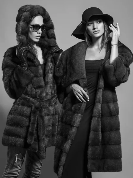 Fashion shot of two elegant beautiful girls (brunette and blonde) in studio on grey background wearing sunglasses, black hat and furs coat . Shopping inspiration. Monochrome (black and white)  photo — Zdjęcie stockowe