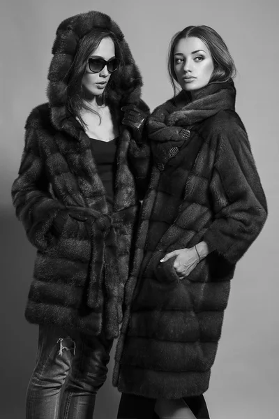 Fashion shot of two elegant beautiful girls (brunette and blonde) in studio on grey background wearing sunglasses and furs coat . Shopping inspiration. Monochrome (black and white)  photo — Stock Fotó