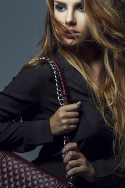 Fashion blonde girl holding Burgundy handbag with long hair, sexy make up and black shirt posing on grey background.