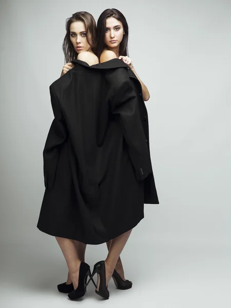 Fashion shot of two elegant beautiful brunette girls  in studio on grey background, dressed in men coat and classic black shoes . Shopping inspiration. — Stock Photo, Image