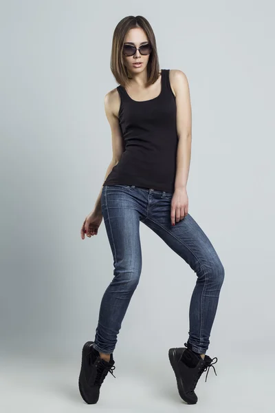 Moderní mladá žena dívka v brýle nosit džíny a černý top pózuje na šedém pozadí. — Stock fotografie