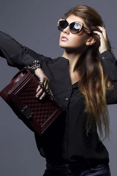 Retrato de una hermosa chica posando en el estudio en camisa negra, vaqueros azules y gafas de sol de moda, sosteniendo bolso de Borgoña. El concepto de las mujeres elegantes y sexy. piel y cuerpo perfectos — Foto de Stock