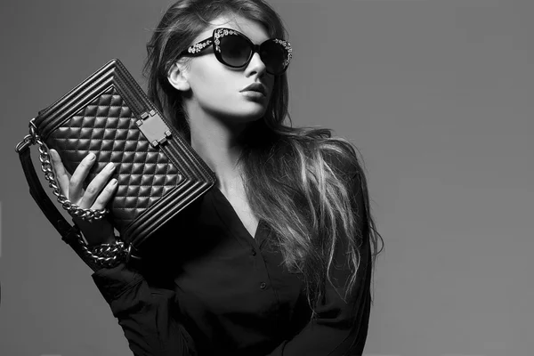 Retrato de una hermosa chica posando en el estudio en camisa negra, vaqueros azules y gafas de sol de moda, sosteniendo bolso de Borgoña. El concepto de las mujeres elegantes y sexy. piel y cuerpo perfectos —  Fotos de Stock