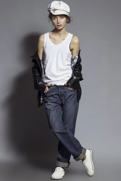 High fashion look. Portrait of a fashionable model with natural make up and perfect skin, dressed in men's jeans, white shirt, black jacket, stylish white hat and sneakers.  Studio shot — Zdjęcie stockowe