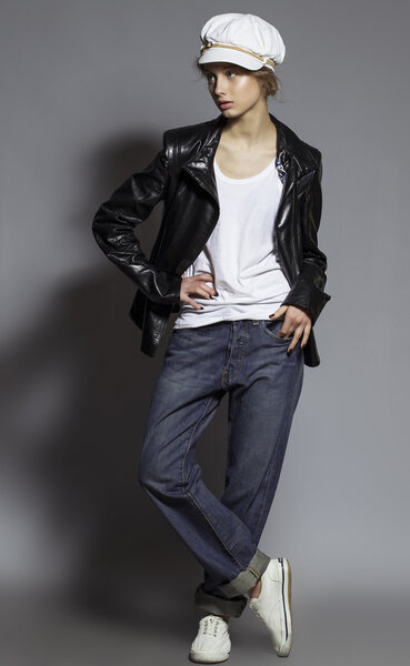 Высокая мода. Портрет модной модели с натуральным макияжем и идеальной кожей, одетый в мужские джинсы, белую рубашку, черную куртку, стильную белую шляпу и кроссовки. Снимок студии
