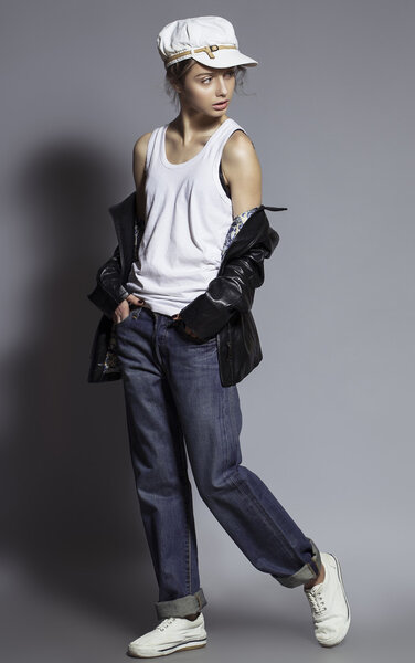 Высокая мода. Портрет модной модели с натуральным макияжем и идеальной кожей, одетый в мужские джинсы, белую рубашку, черную куртку, стильную белую шляпу и кроссовки. Снимок студии
