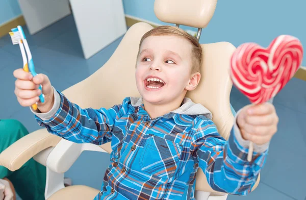 Niño muy feliz después de que el médico en la clínica, sosteniendo un caramelo del corazón y cepillos de dientes, y hermosas sonrisas — Foto de Stock