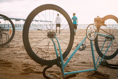 Bisiklet plajın ön tarafındaki kumsalda baş aşağı duruyor ve sırtlarında genç insanlar denize doğru güneşin doğuşunu seyrediyor. Genç insanlar sahilde boş vakitlerinin tadını çıkarıyorlar. Telif alanı olan bir arka plan. Spor anlayışı. Açık hava konsepti. Boş 