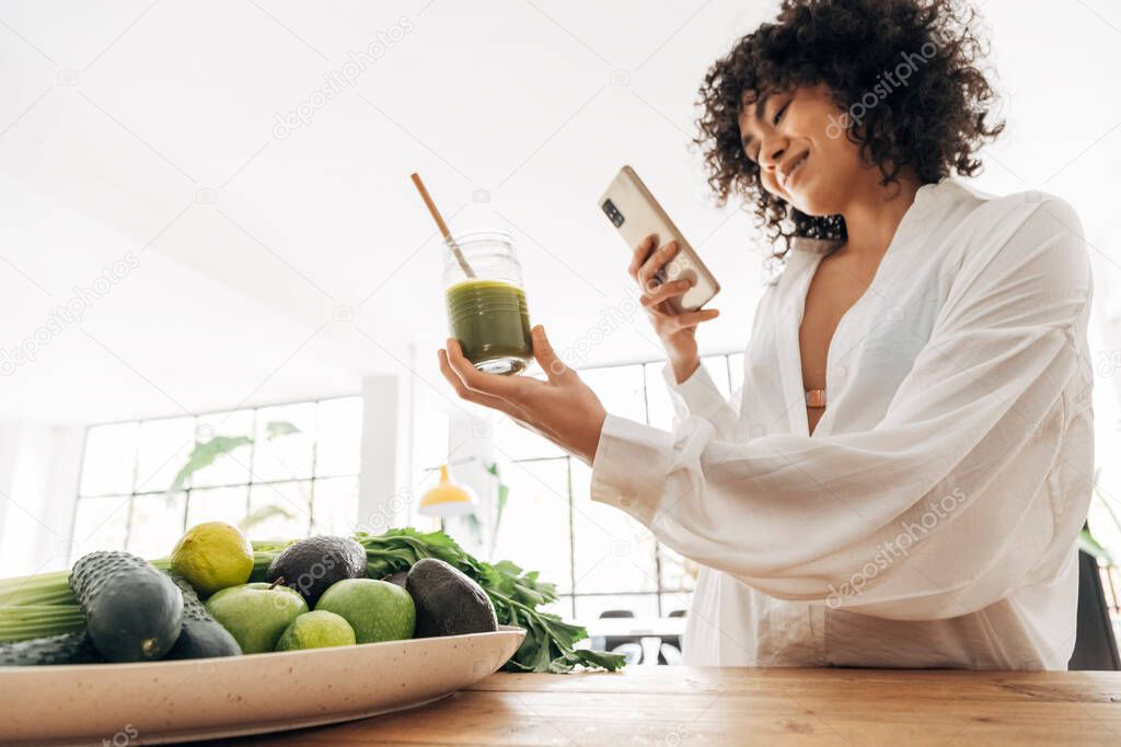 Young african american woman taking photo of green juice and vegetables with cellphone at home. Home concept. Technology concept. Health concept. Copy space