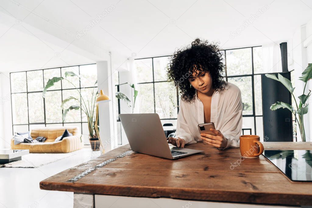 Young african american woman checking cellphone and using laptop in kitchen counter in bright loft apartment. Technology concept. Work from home concept. Copy space