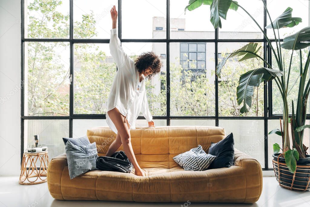 Beautiful African american young woman dancing on couch, having fun, carefree in a big, bright loft apartment. Home concept. Freedom concept. Fun concept.