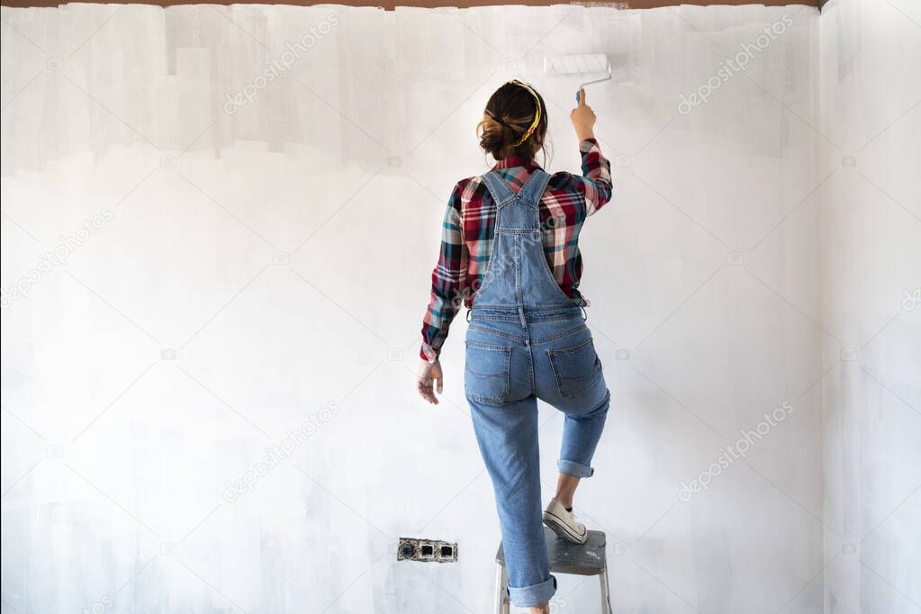 Young woman on a ladder painting new apartment walls in color white with paint roller. Renovating house. Real estate concept. Copy space