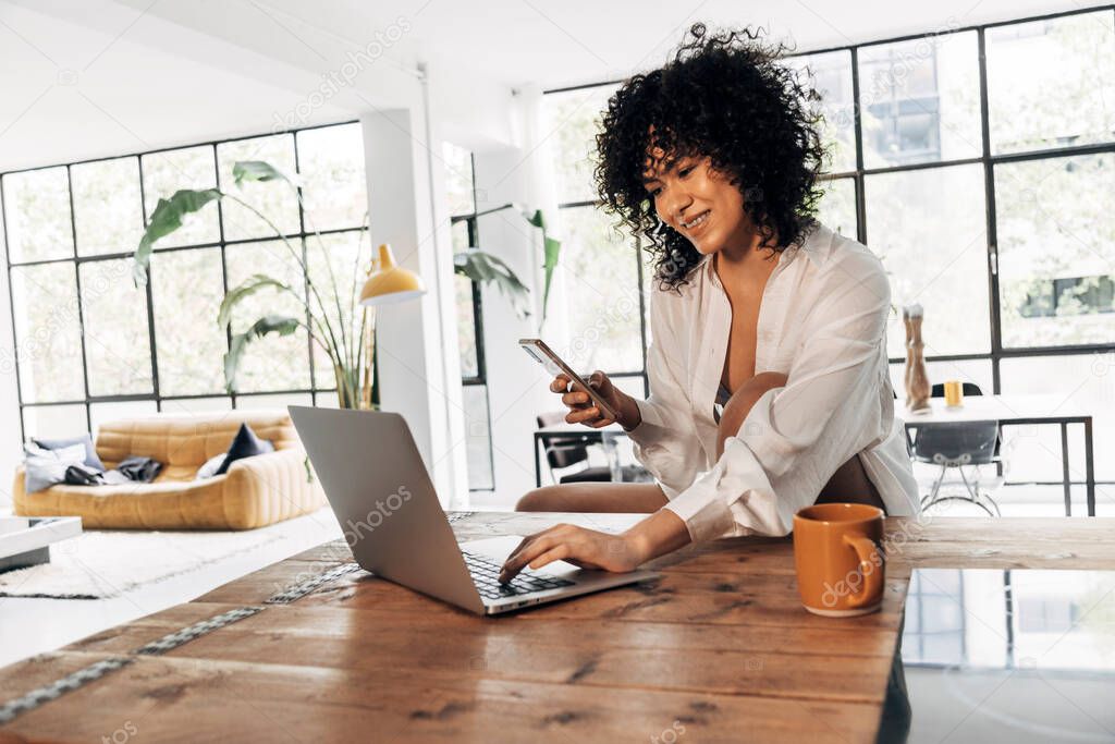 Young beautiful african american woman typing on laptop on kitchen counter, having coffee and holding cellphone. Copy space. Home concept. Technology concept.
