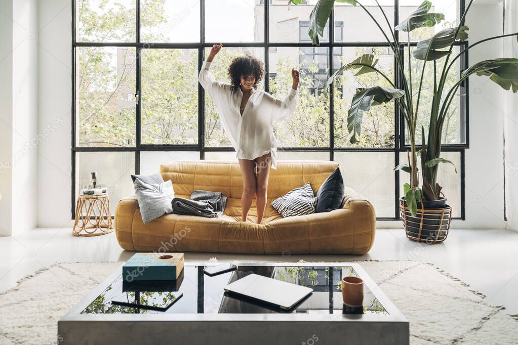 Young multiracial woman dancing to music on the couch in a bright living room. Home concept.