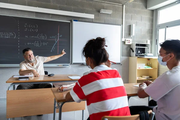 Mature white man teacher points blackboard in high school classroom. Multiracial teen boy and girl listen to explanation. Copy space. Education concept.