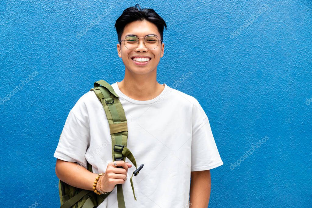 Portrait of happy and smiling teen asian boy high school student looking at camera with backpack on blue background. Copy space.