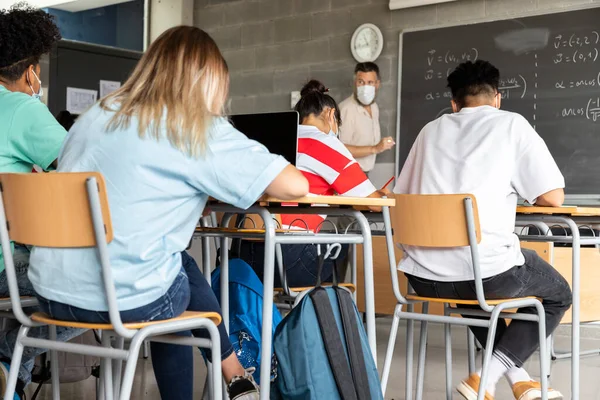 Mathematics class. Mature white male teacher wearing face mask in classroom with group of multiracial teen high school students. Education concept. Healthcare concept.