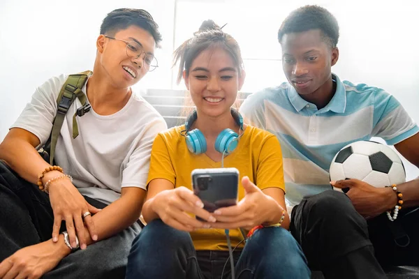 Multiracial group of teen high school classmates sit on stairs looking at teenage girl cell phone. Back to school concept. Technology addiction concept.