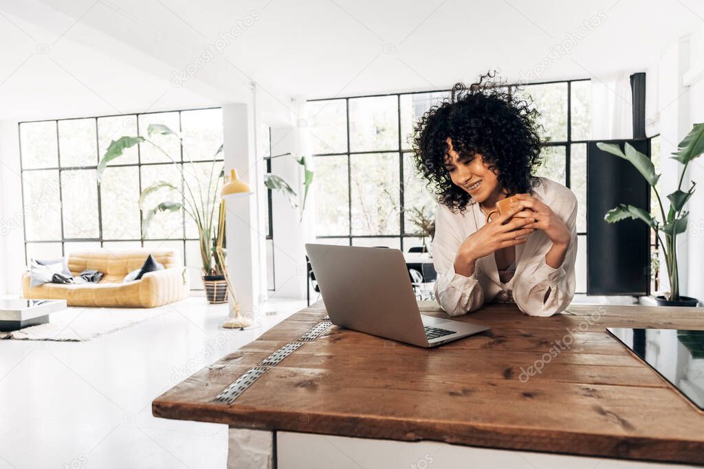 Young african american woman having a coffee in the morning looking laptop in bright loft kitchen. Home concept. Technology concept. Copy space